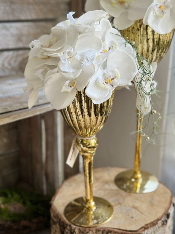 Kompozycja w wysokim szklanym złotym kielichu. Dekoracja została wykonana ze silikonowymi kwiatami storczyka.