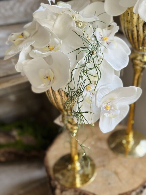 Kompozycja w wysokim szklanym złotym kielichu. Dekoracja została wykonana ze silikonowymi kwiatami storczyka.