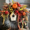 Jesienny wianek wykonany z pomarańczowych oraz brązowych liści. Całość dopełniona dyniami, owocami, kwiatami oraz drewnianym domkiem.