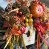 Jesienny wianek wykonany z pomarańczowych oraz brązowych liści. Całość dopełniona dyniami, owocami, kwiatami oraz drewnianym domkiem.