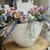 Kompozycja jesienna wykonana w białej szkliwionej ceramicznej osłonce z dodatkiem wrzosów i kwiatów. Całość została dopełniona biało-srebrnym ceramicznym domkiem .