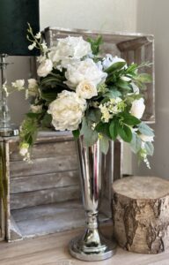 Kompozycja wykonana w srebrnym metalowym wazonie. W kompozycji zostały wykorzystane biało-kremowe kwiaty hortensji, peoni, róży i storczyka.