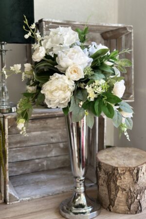 Kompozycja wykonana w srebrnym metalowym wazonie. W kompozycji zostały wykorzystane biało-kremowe kwiaty hortensji, peoni, róży i storczyka.