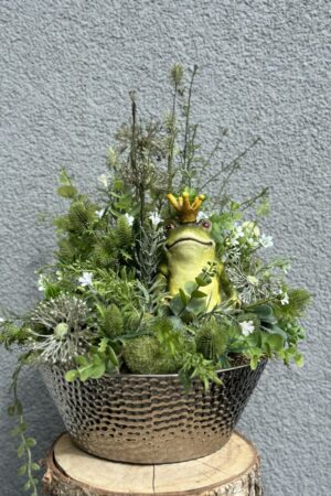 Kompozycja z kwiatów sztucznych z ceramiczną zieloną żabą w srebrnym ceramicznym naczyniu.