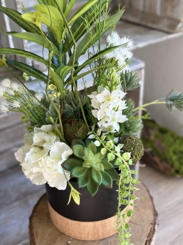 Kompozycja sztuczna wykonana z zielonych roślin draceny, sukulentów, białej hortensji. Dekoracja została osadzona ceramicznej osłonce w kolorze czarno-drewnianym