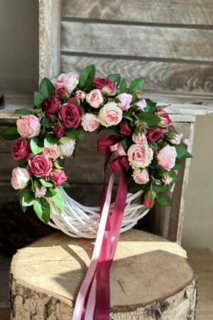 Wianek wykonany na białym wiklinowym kole z dodatkiem kwiatów róży gałązkowej w kolorze jasno różowym i amarantowym.
