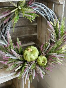 Jesienny wianek wykonany na wiklinowym szarym kole z dodatkiem wrzosów fioletowych, białych i zielonych. Całość została upiększona aksamitnymi zielonymi dyniami.