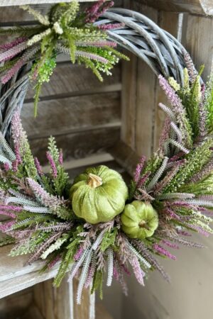 Jesienny wianek wykonany na wiklinowym szarym kole z dodatkiem wrzosów fioletowych, białych i zielonych. Całość została upiększona aksamitnymi zielonymi dyniami.