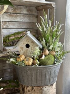 Kompozycja jesienna wykonana w metalowym naczyniu z drewnianym domku. Dekoracja została uzupełniona dyniami, ptaszkiem i sztucznym mchem.