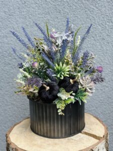 Jesienna kompozycja w odcieniach błękitu i fioletu dopełniona czarnymi aksamitnymi dyniami. Dekoracja została wykonana w czarnej matowej osłonce.