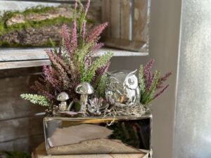 Kompozycja jesienna w srebrnym naczyniu z ceramiczną sową i grzybkami. Całość uzupełniona wrzosami w kolorze fioletowym i zielonym.